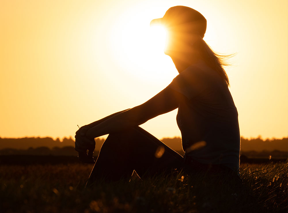 A silhouette of a woman wearing a ballcap, sitting in a field at sunset.